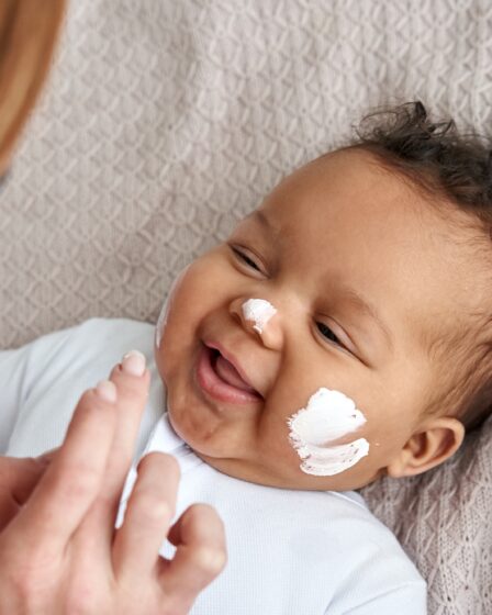 Kaukaska matka nakłada krem ​​nawilżający do pielęgnacji zdrowej skóry na uroczą, zabawną, afroamerykańską twarz córki dziewczynki.